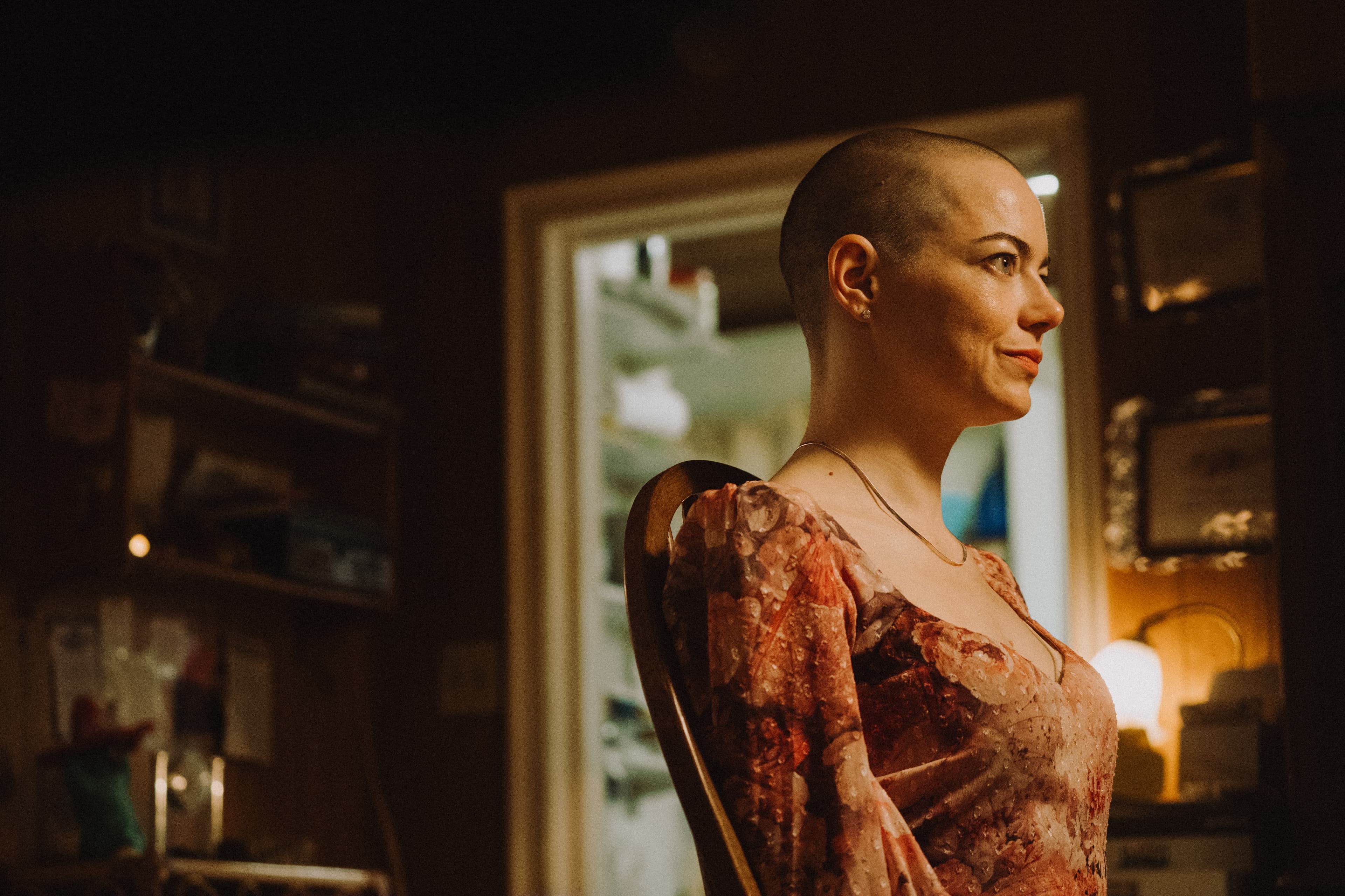 Emma Stone sits in a basement with her head shaved and wearing a dated floral dress in a still from "Bugonia." 