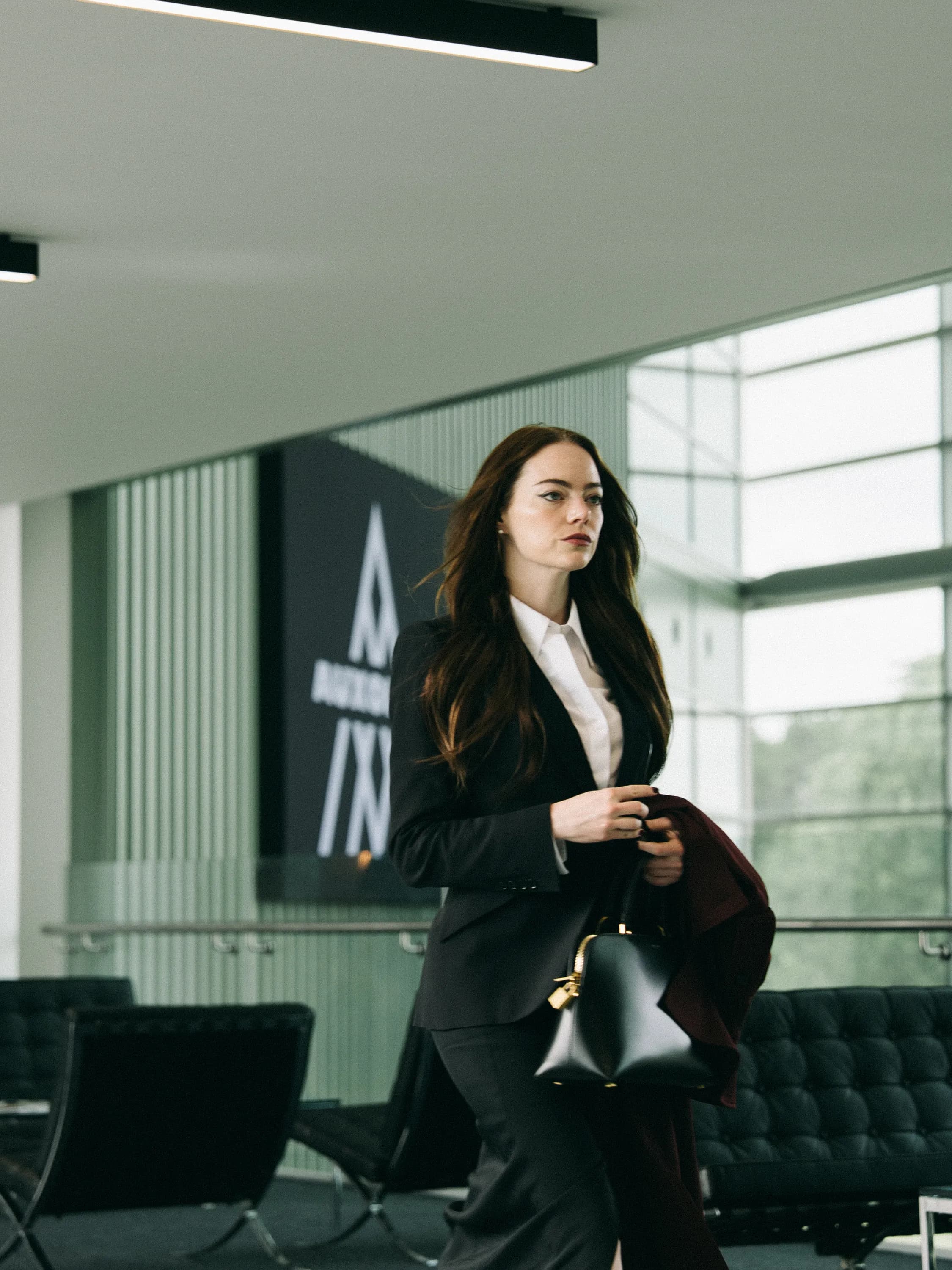 Actor Emma Stone stands in a modern office wearing a suit and carrying a black bag in a still from "Bugonia." 