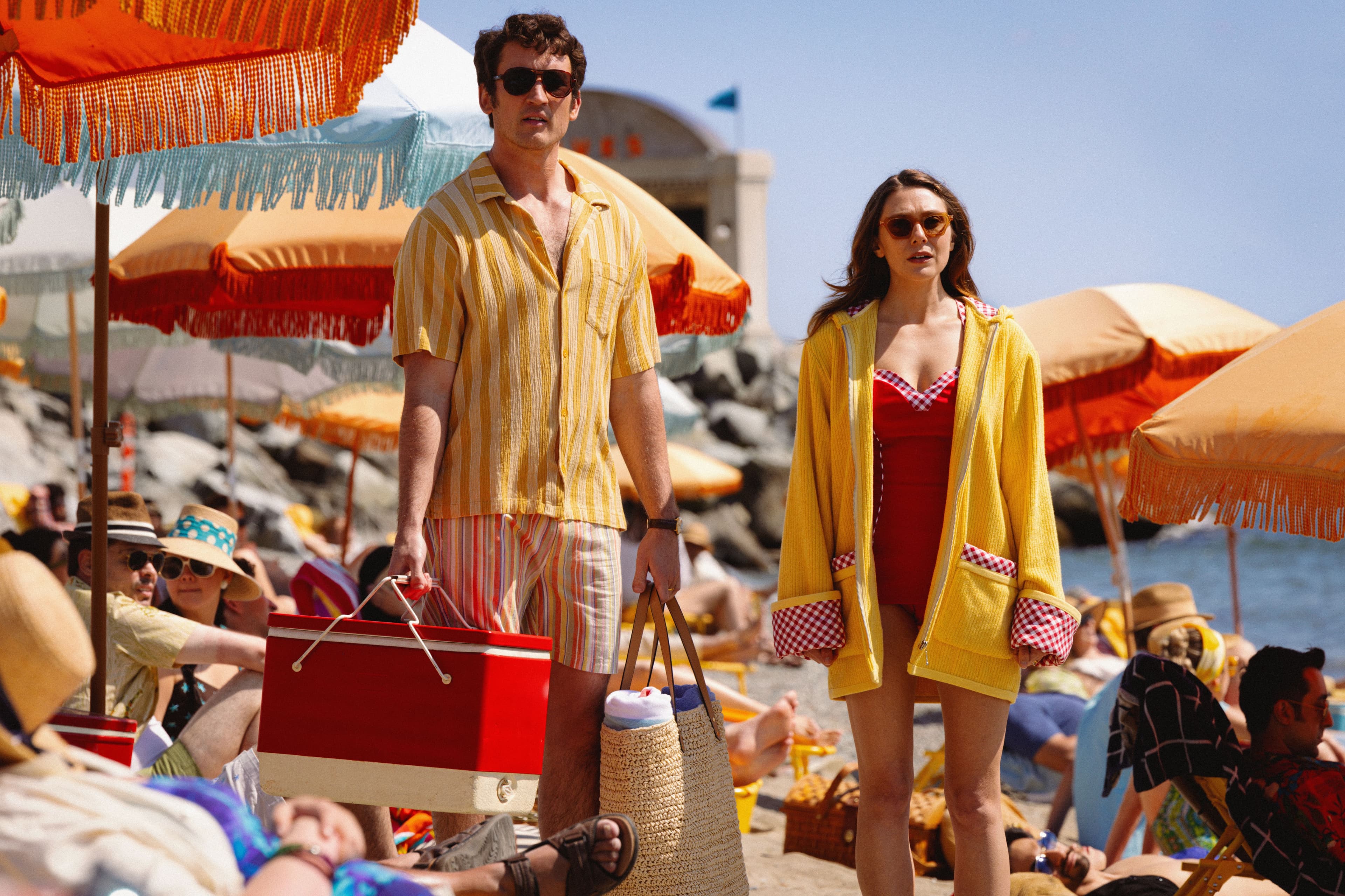 Actors Miles Teller and Elizabeth Olsen stand next to each other on a crowded beach in a still from Eternity. 