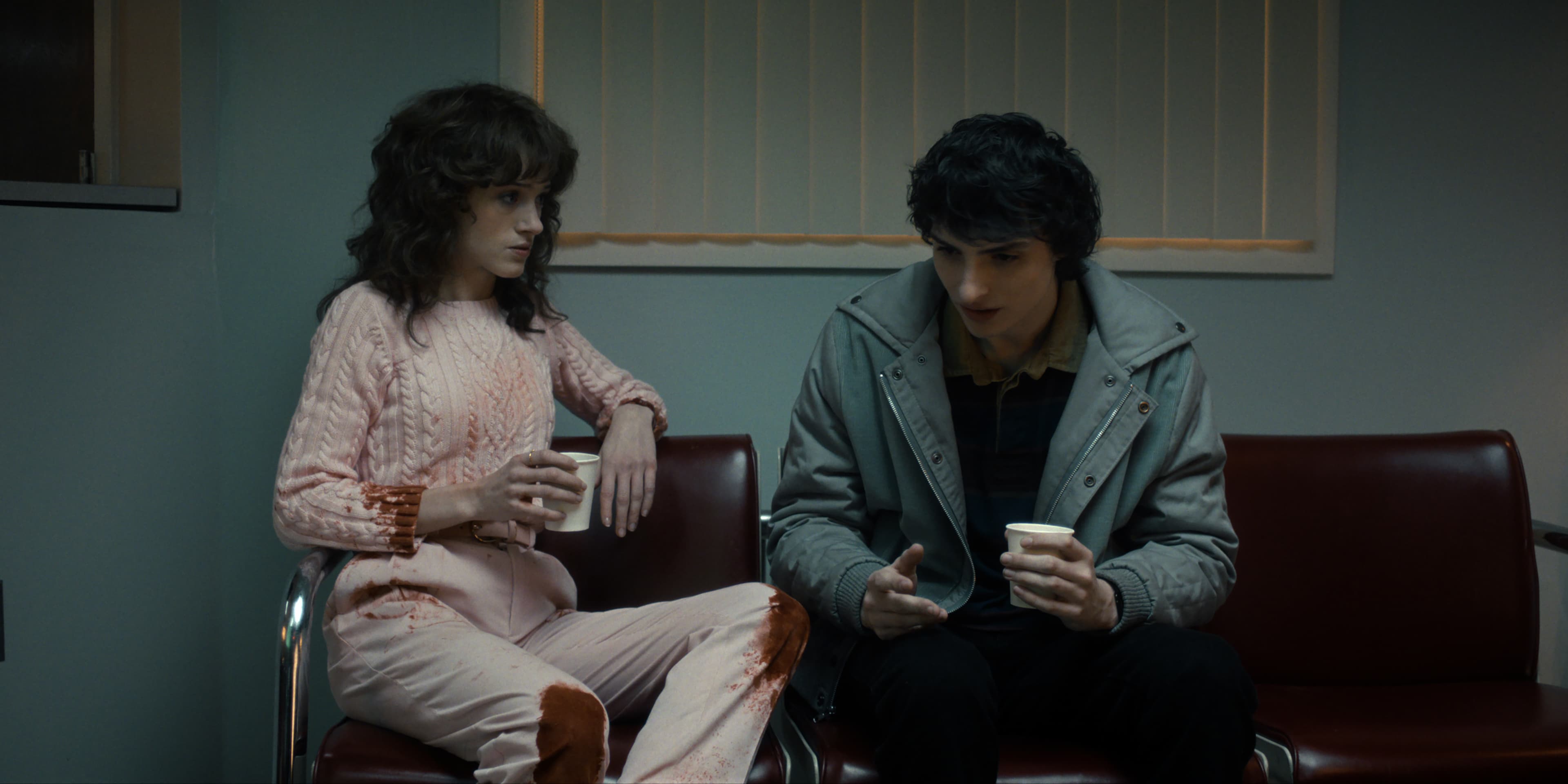 Actors Natalia Dyer and Finn Wolfhard sit next to each other in a hospital lobby in a still from Stranger Things season five. 