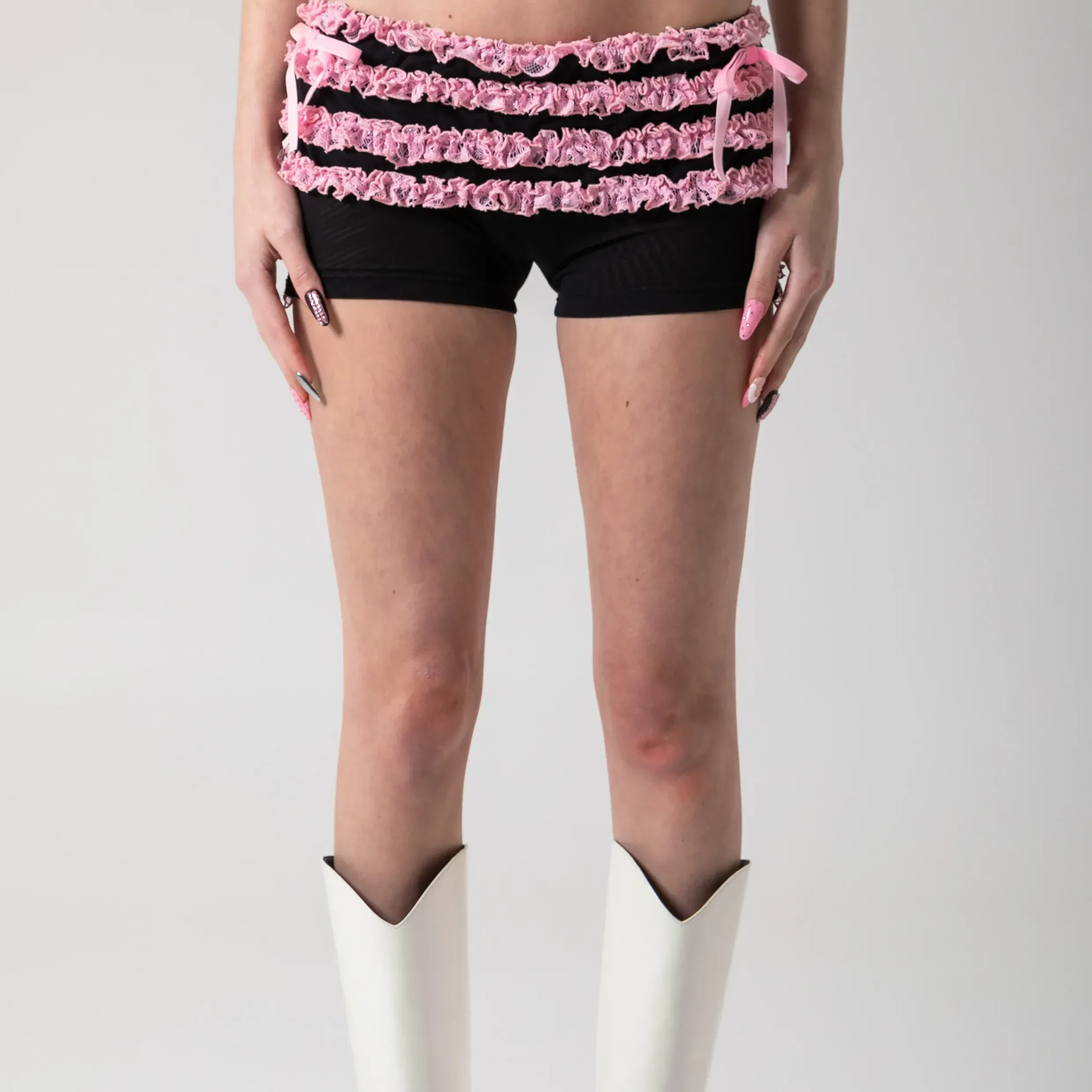 A model wearing black and pink ruffle shorts and white cowboy boots against a gray background