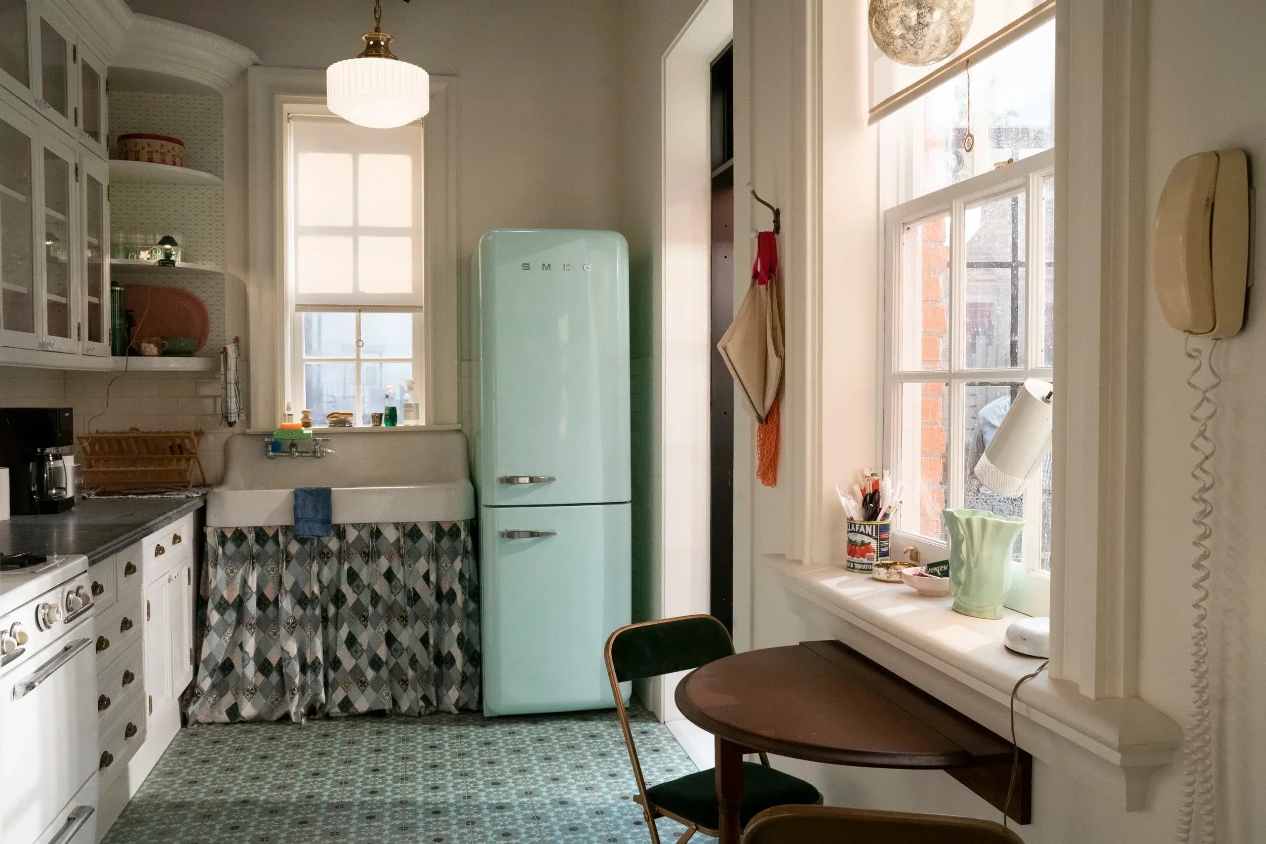 A photo of a kitchen, looking at the seafoam vintage-stle fridge, small halfmoon shaped wooden table, and window from the show "And Just Like That."