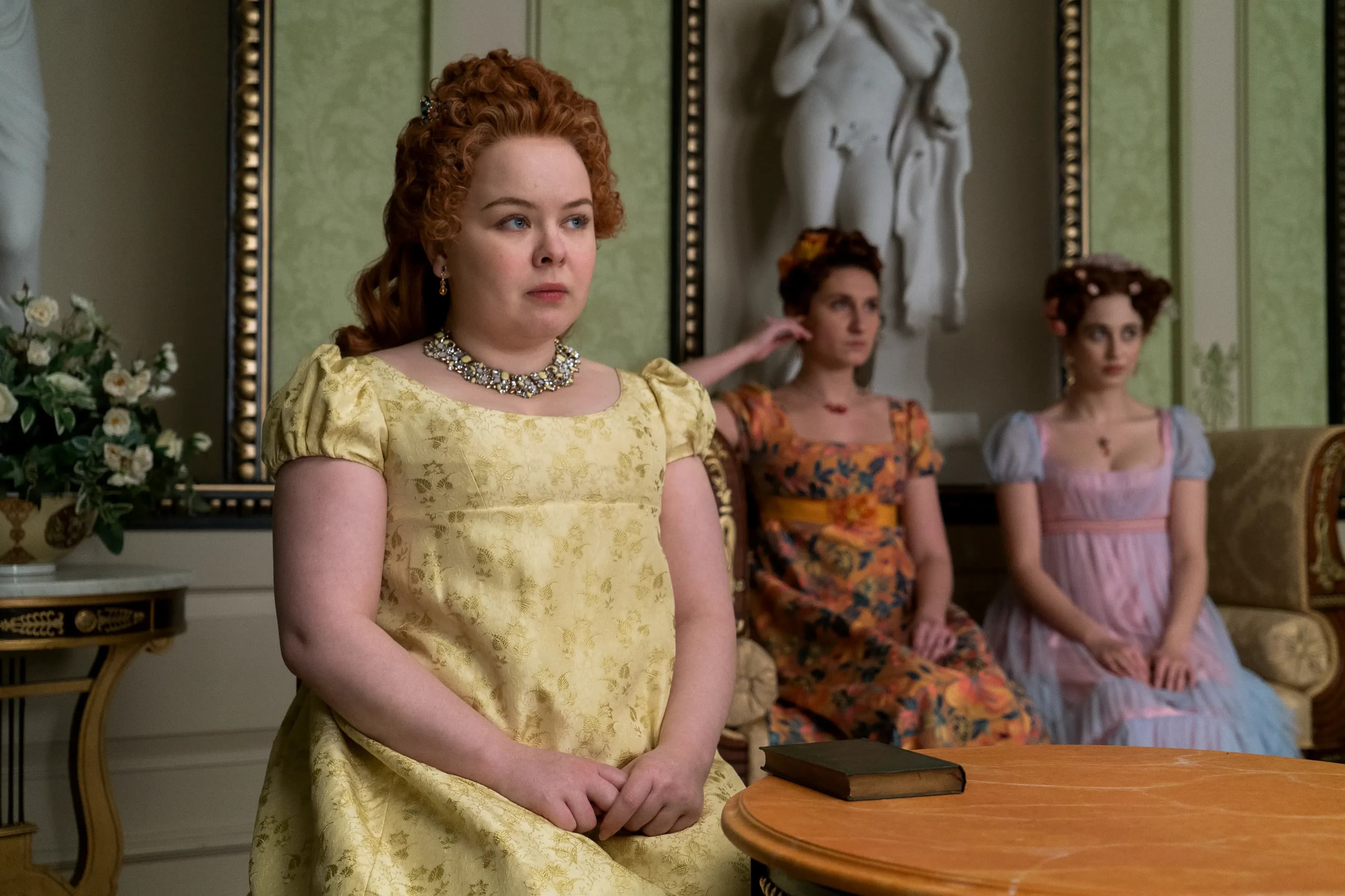 Nicola Coughlan sits at a table with her hands folded, wearing a pale yellow dress in a scene from Bridgerton.