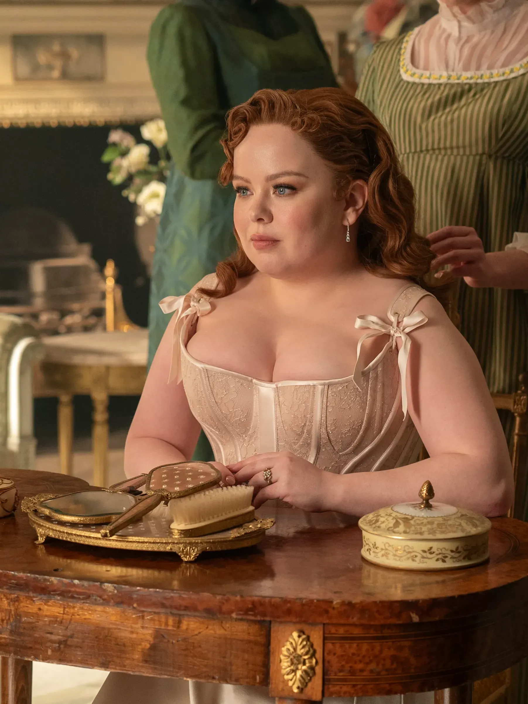 Nicola Coughlan sits at a table with her arms folded, wearing a beige corset dress.