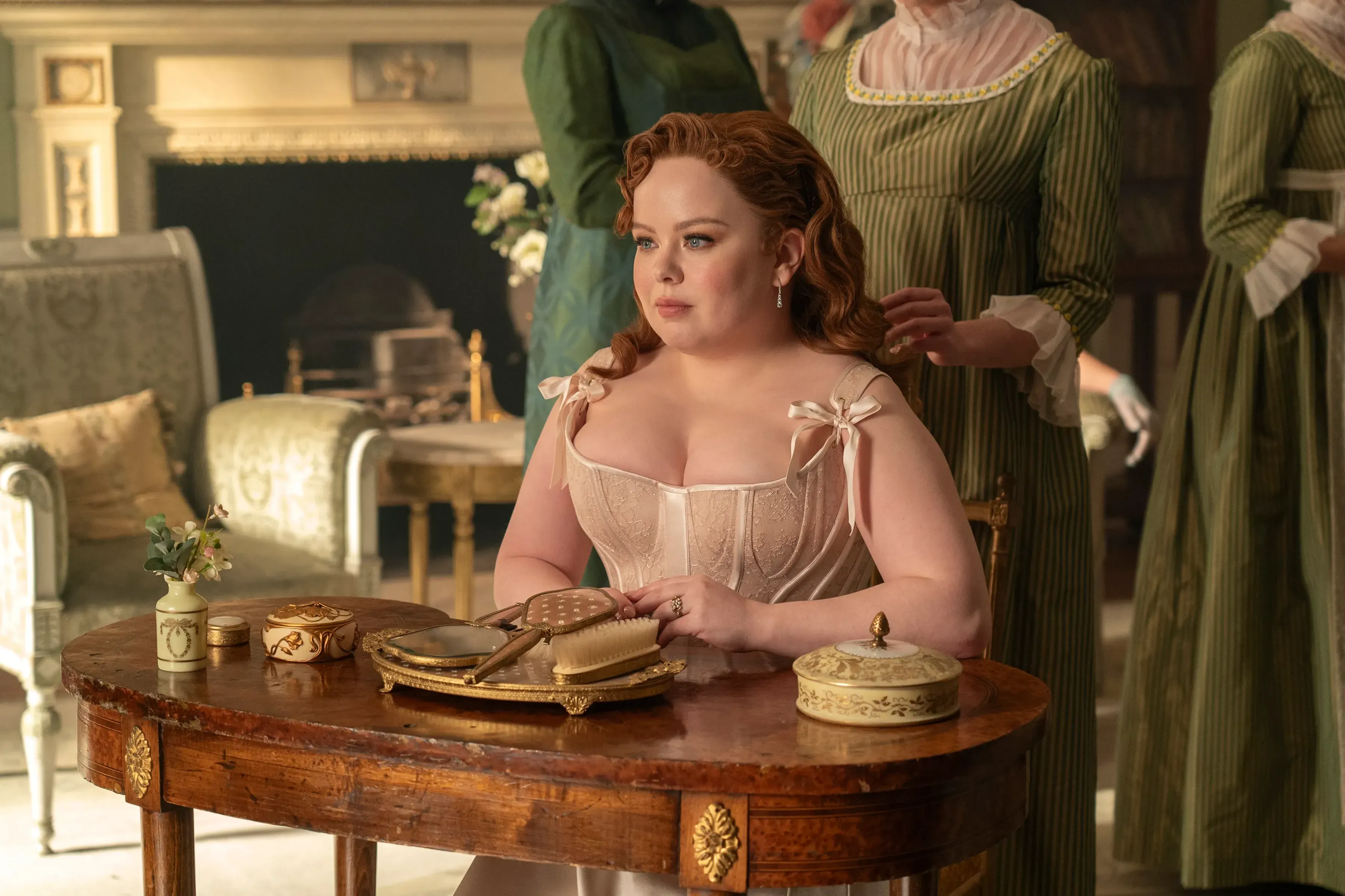 Nicola Coughlan sits at a table with her arms folded, wearing a beige corset dress.
