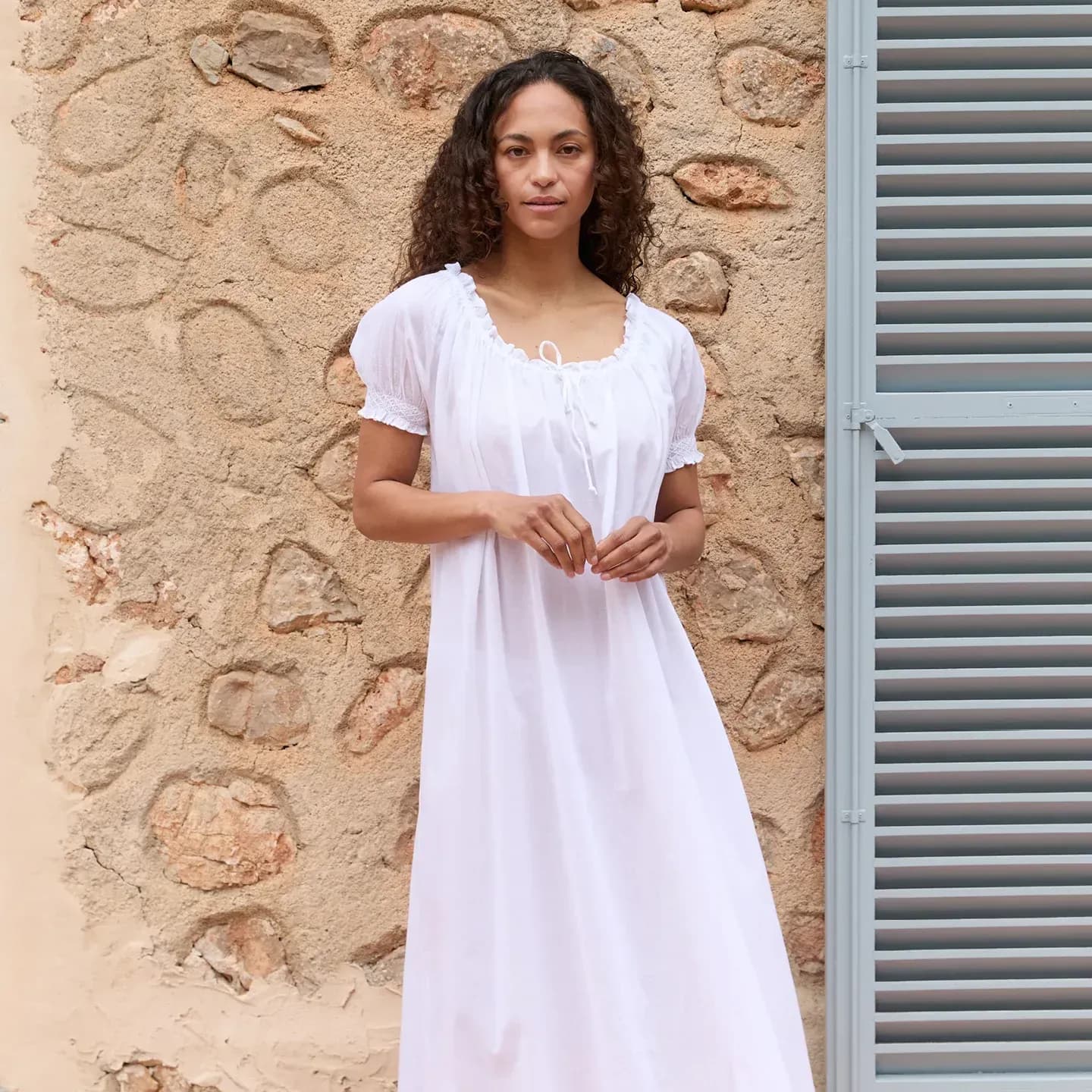 Model standing against a stone wall in a long white nightgown